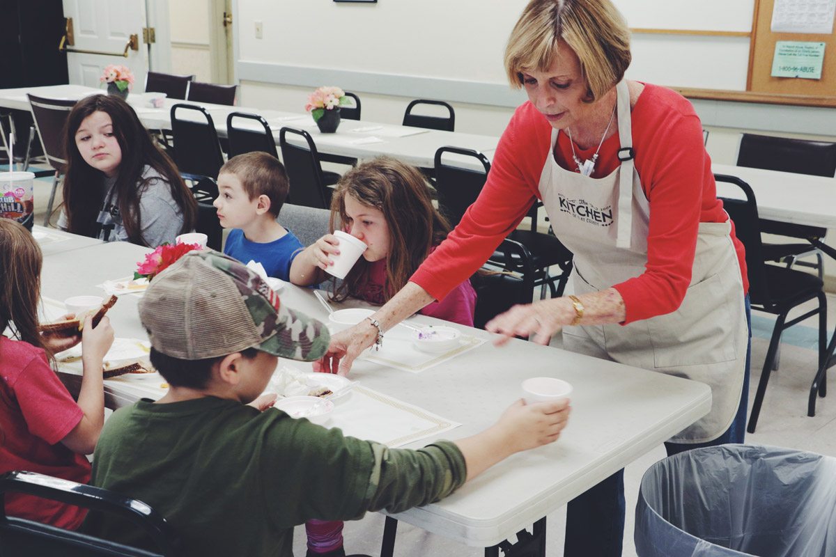 A volunteer in a Kitchen apron serving children their meals at the table