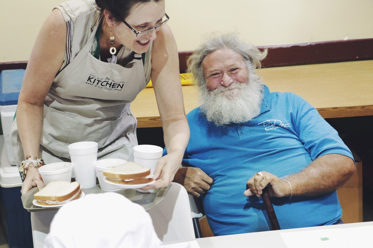A volunteer in a Kitchen of Clay County apron serving soup and sandwiches to a smiling guest